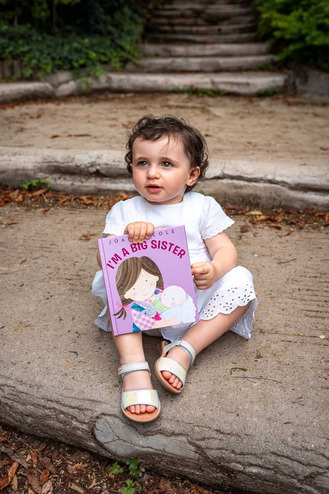 Jeune enfant assise sur des marches en pierre, tenant un livre intitulé 'I'm a Big Sister'
