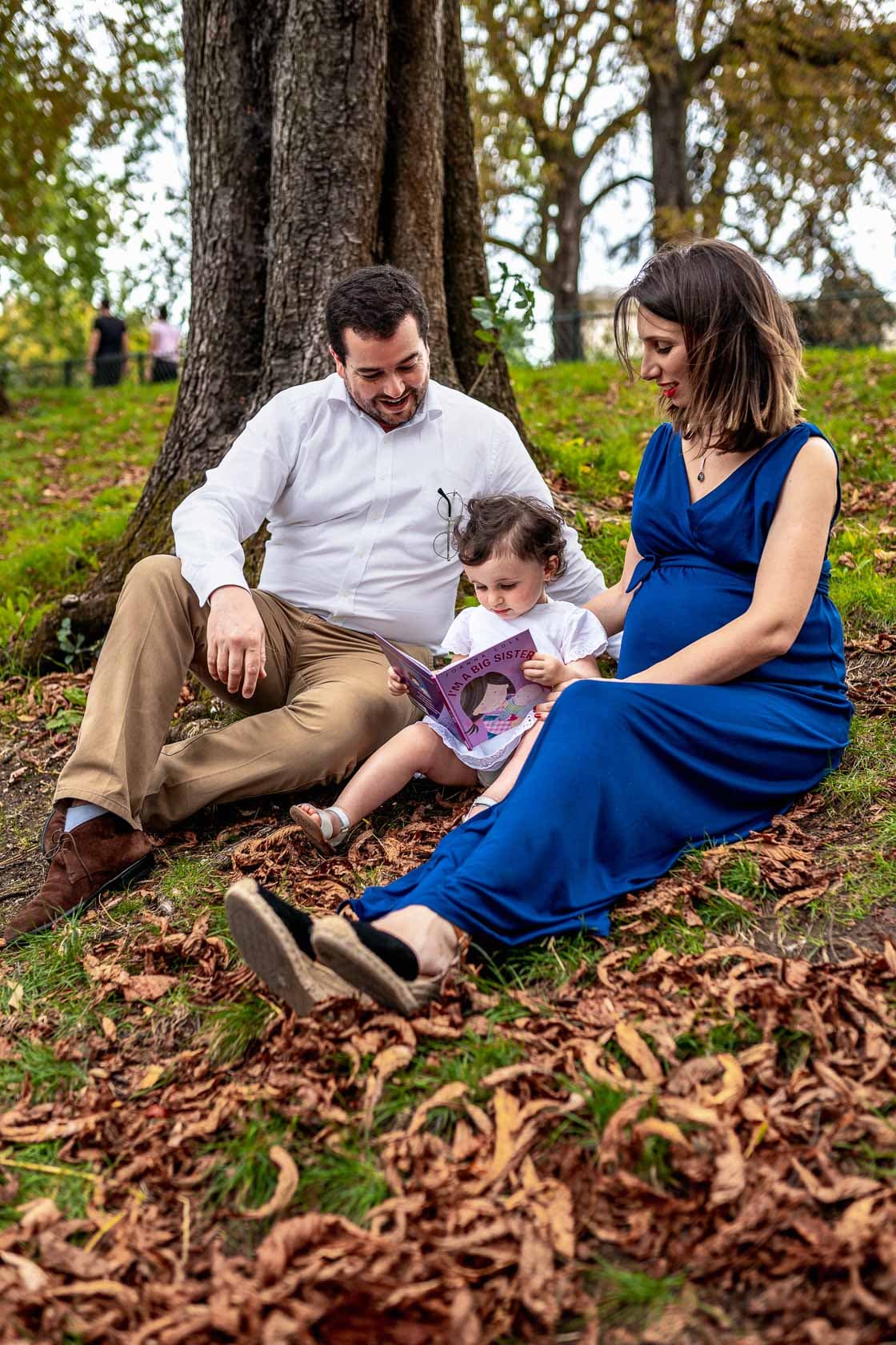 Famille de trois personnes assise au pied d'un grand arbre dans un parc automnal, femme enceinte en robe bleue lisant un livre à une jeune enfant, homme souriant regardant sa fille
