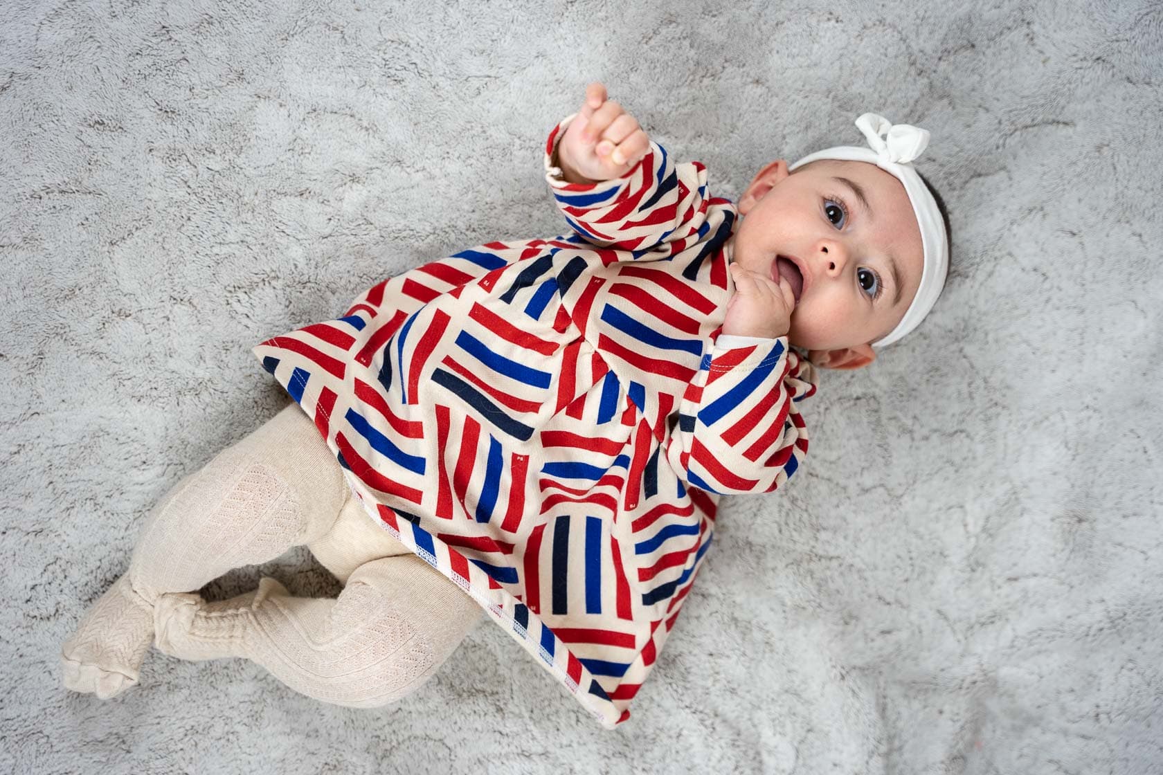 Bébé allongé sur le dos sur une couverture grise, portant une robe à rayures rouges, blanches et bleues et un bandeau blanc