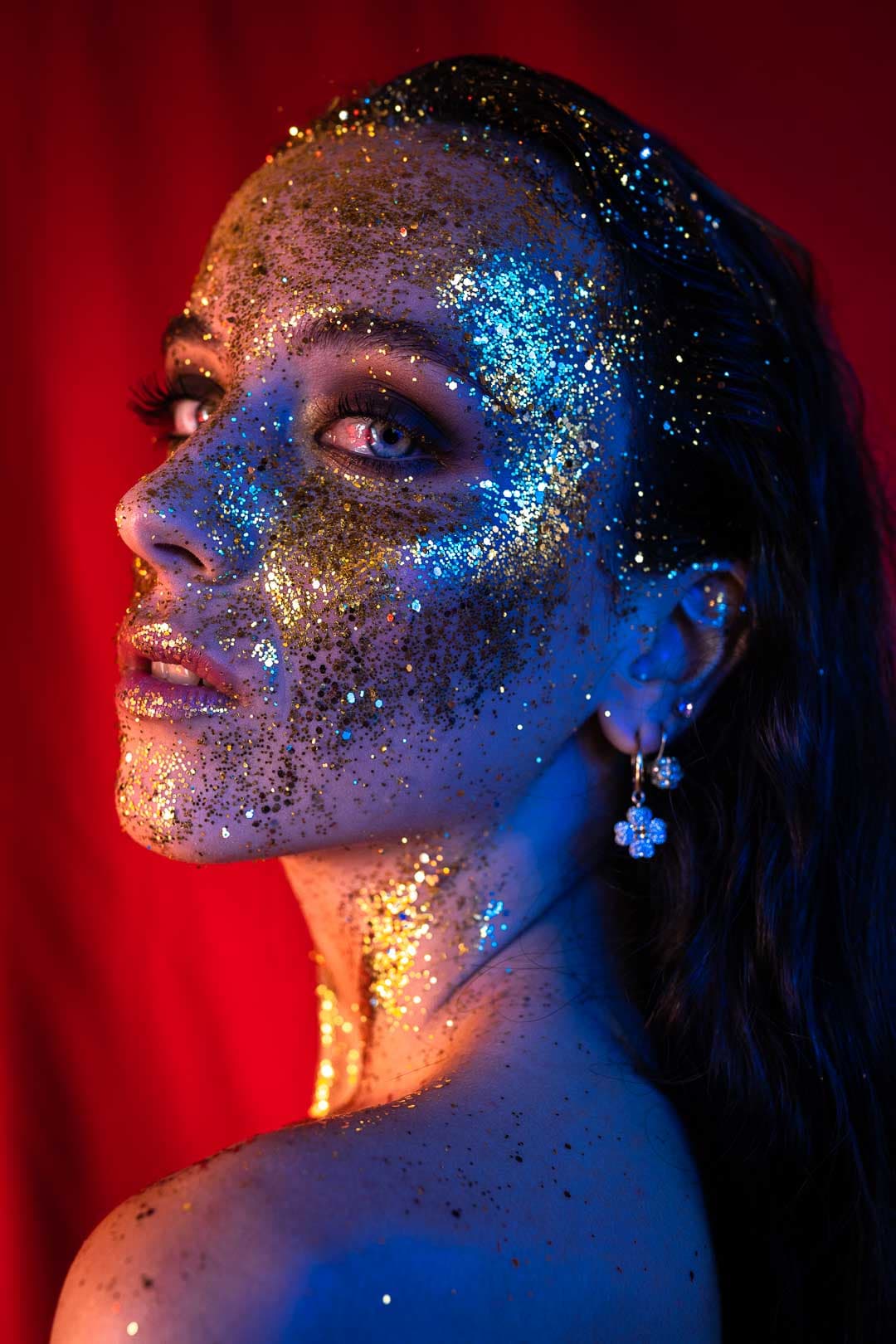 Portrait d'une jeune femme avec le visage couvert de paillettes dorées sous un éclairage rouge et bleu
