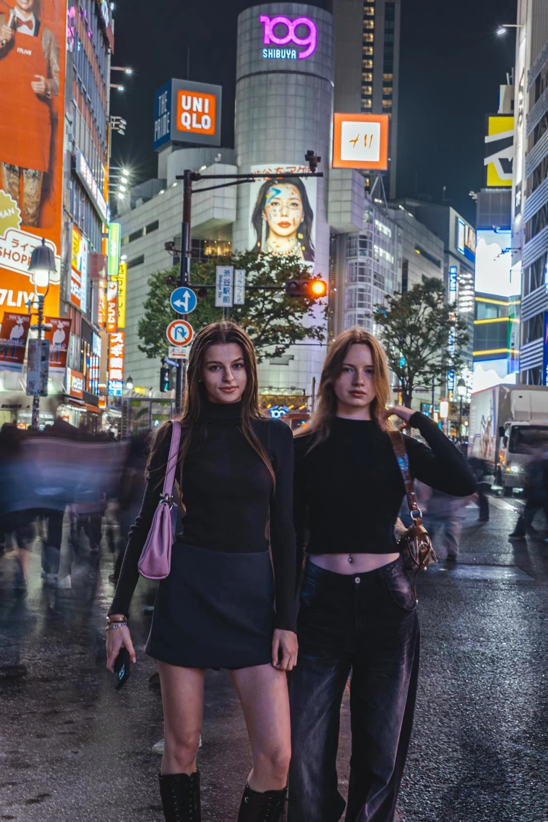 Two friends dressed in black, skirts and wide-legged trousers, at the Shibuya intersection at night
