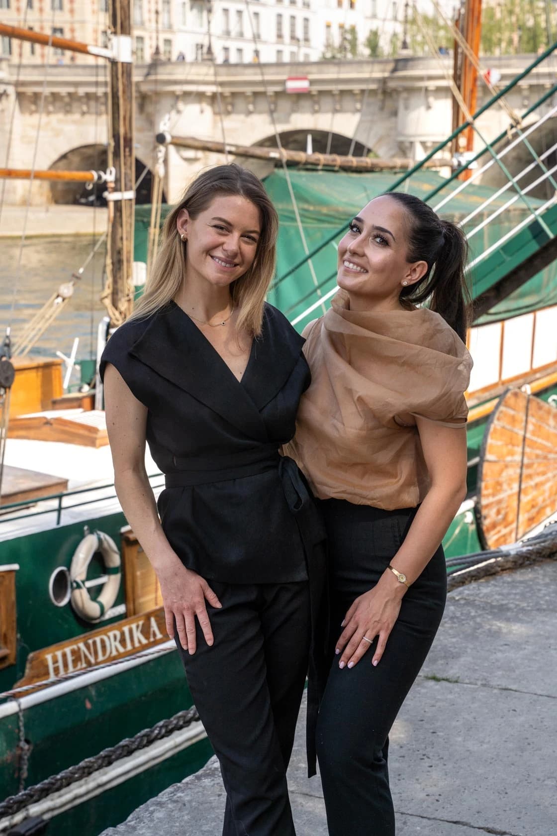 Two smiling friends in black and beige outfits, on the banks of the Seine in front of an old sailboat, the Hendrika, Paris