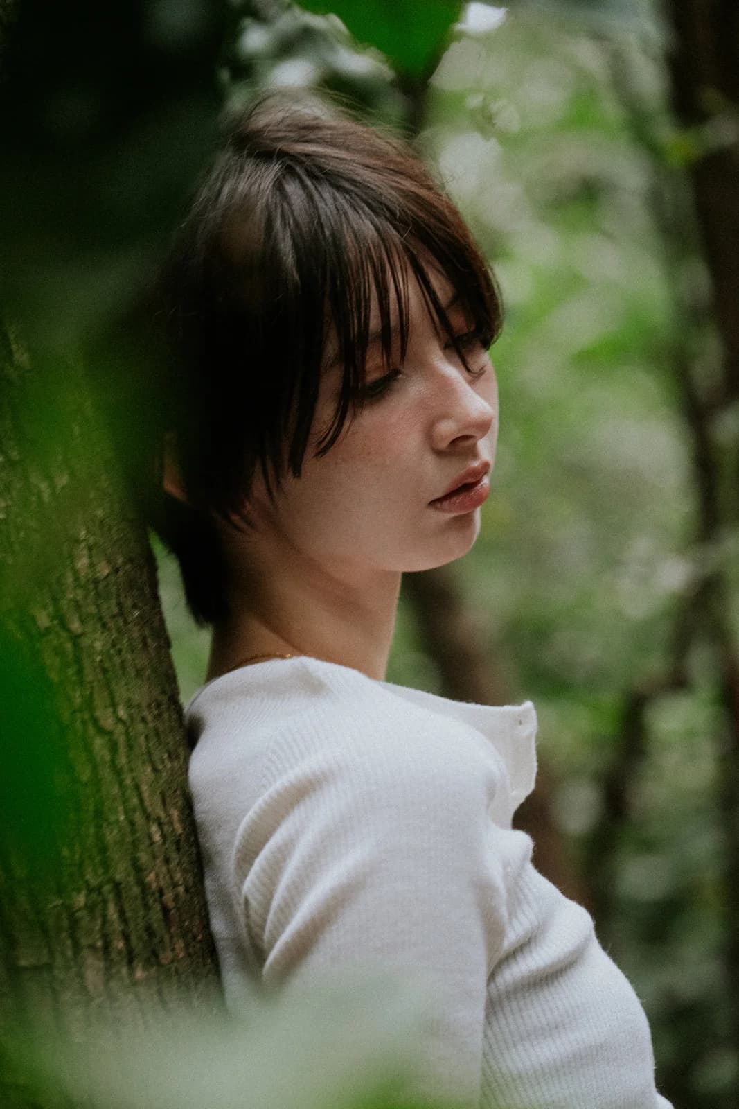 Portrait de profil d'une femme brune aux cheveux courts, appuyée contre un arbre entouré de fleurs blanches, lumière douce
