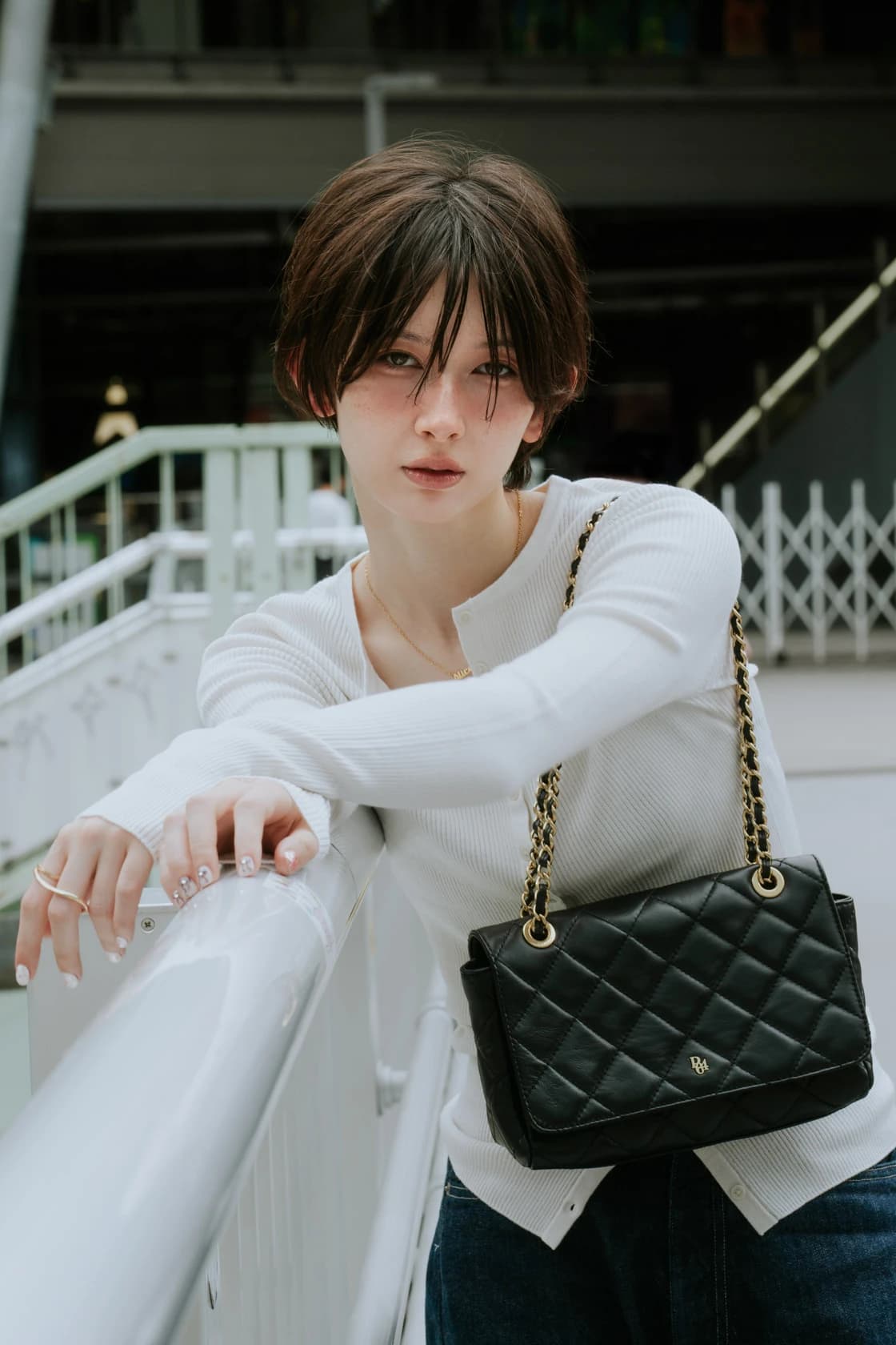 Femme brune aux cheveux courts, pull blanc côtelé, sac matelassé noir à chaîne dorée, appuyée sur une rambarde, plan rapproché à Tokyo