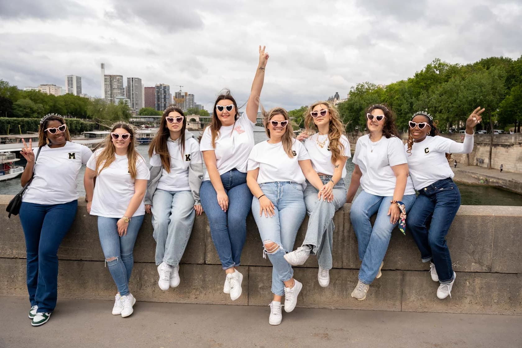 A group of eight friends wearing heart-shaped glasses sitting on a low wall by the Seine, with the Paris skyline in the background.