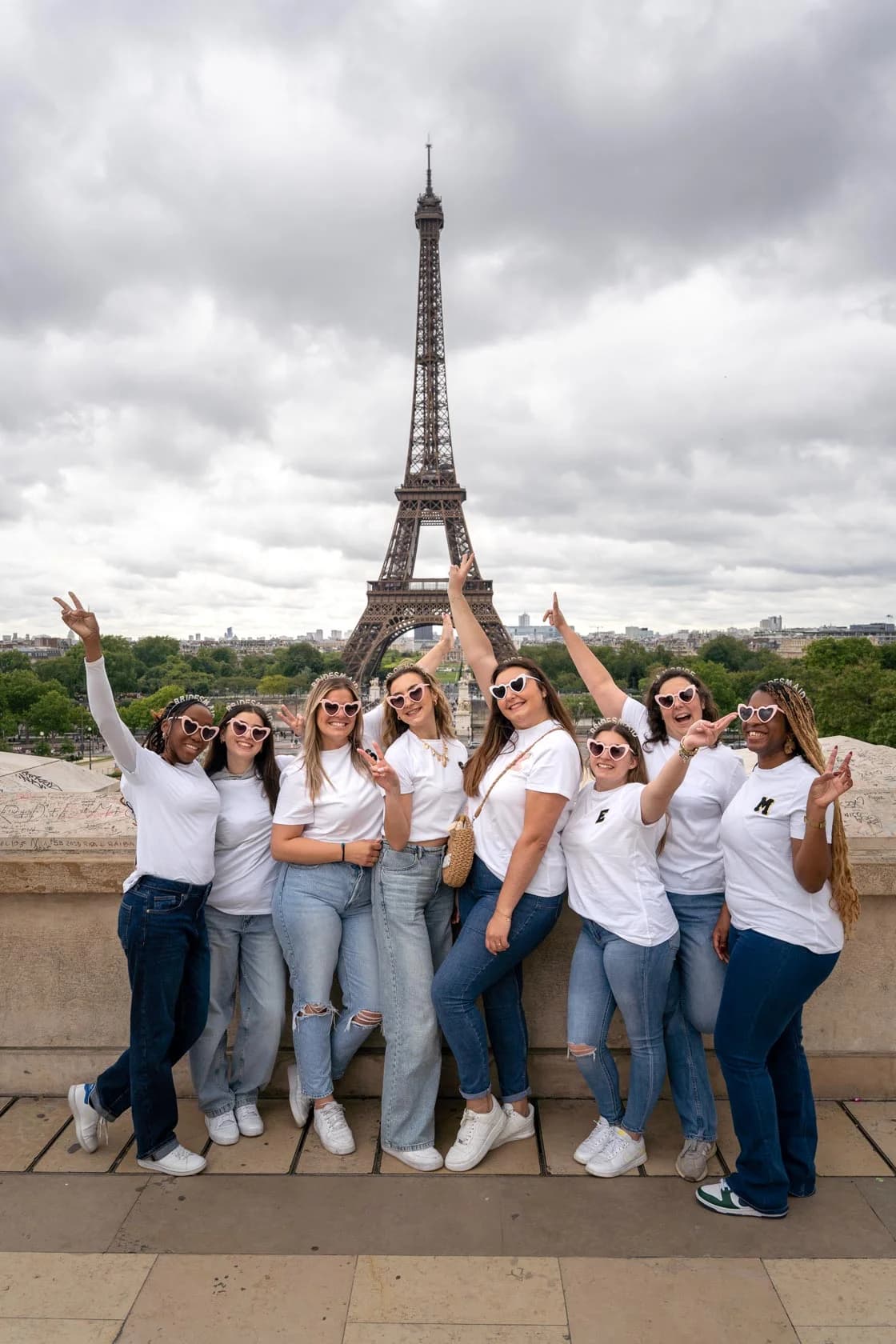 A group of eight friends in white t-shirts and heart-shaped glasses, arms raised, on the Trocadero plaza facing the Eiffel Tower