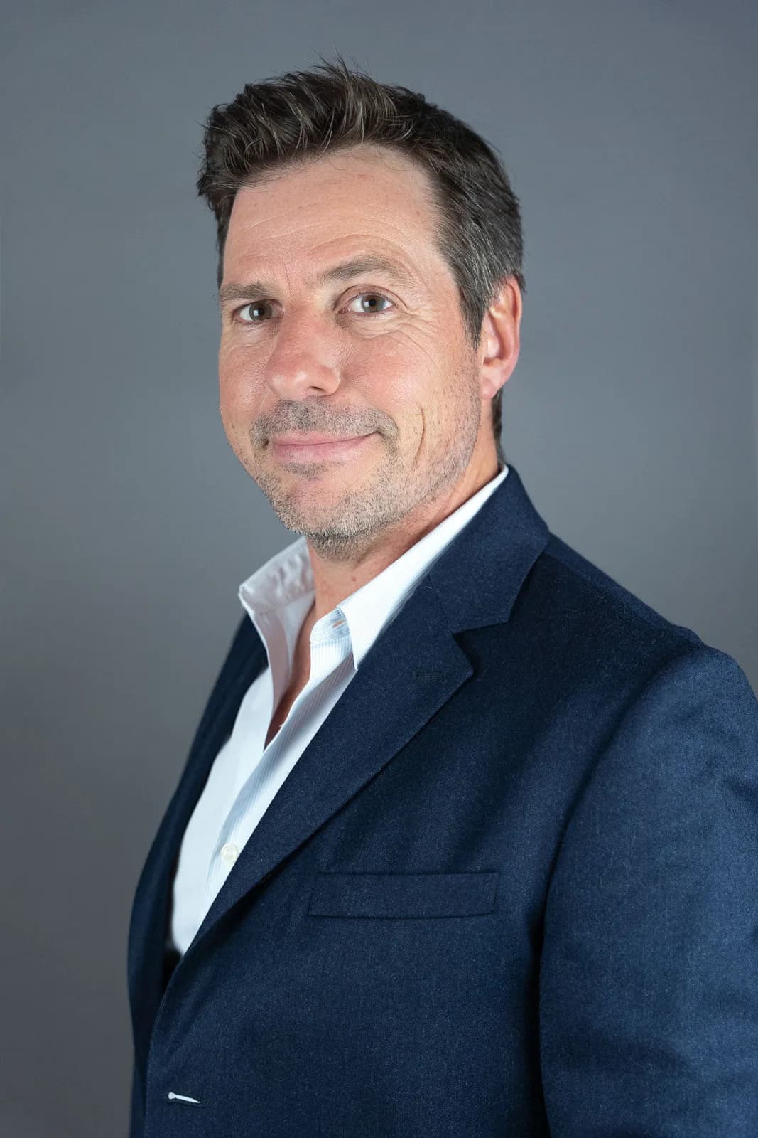 Portrait d'un homme souriant avec des cheveux poivre et sel, portant un blazer bleu sur une chemise blanche ouverte