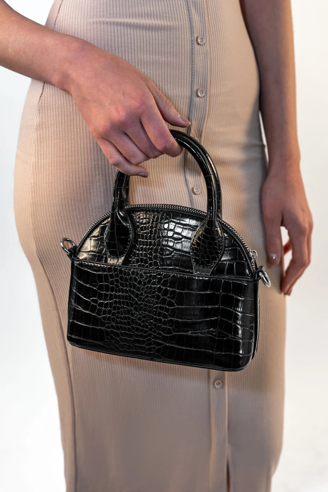 Gros plan sur un sac à main noir façon croco tenu à la main, robe beige boutonnée en arrière-plan