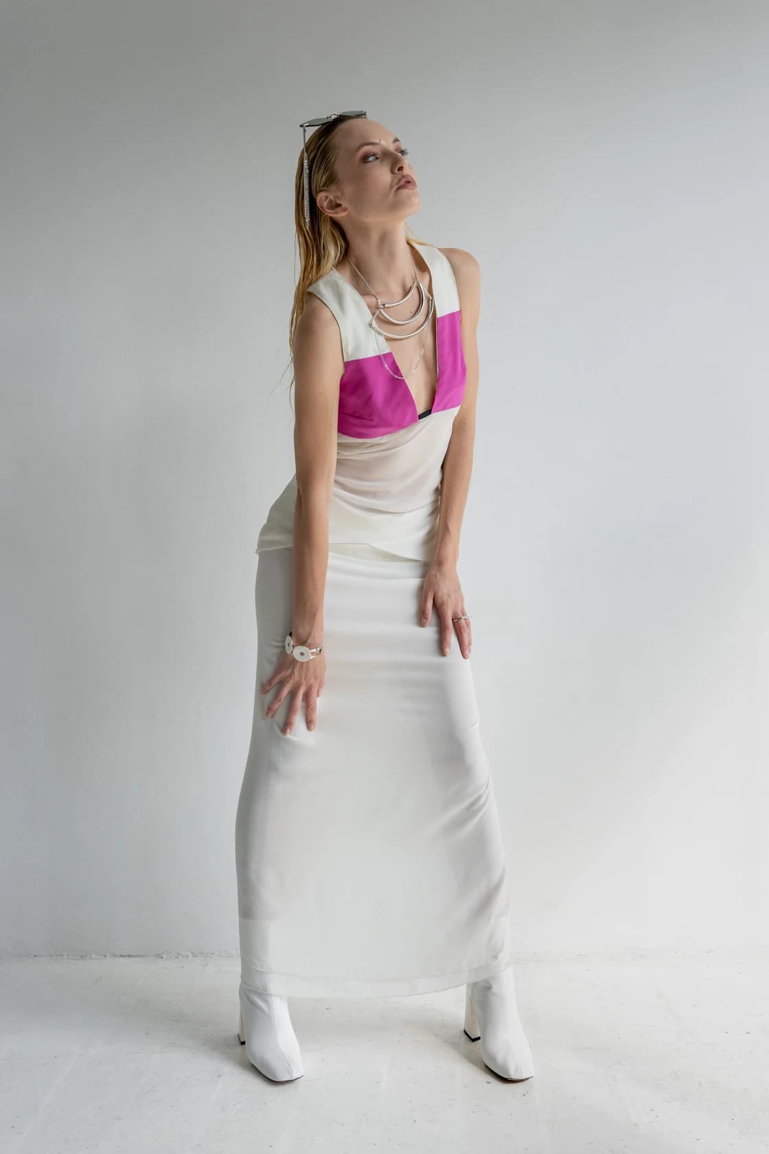 Mannequin en robe blanche à empiècement fuchsia, lunettes de soleil sur la tête, colliers superposés, bottes blanches, studio