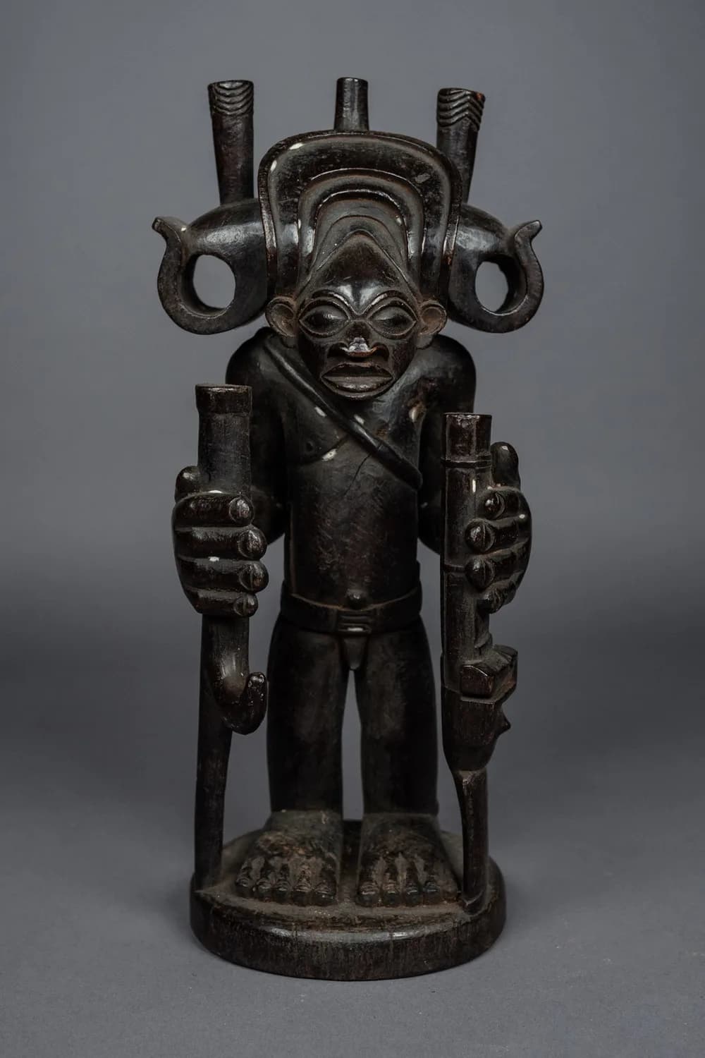 African statue in dark wood depicting a figure holding two long pipes, on a grey background