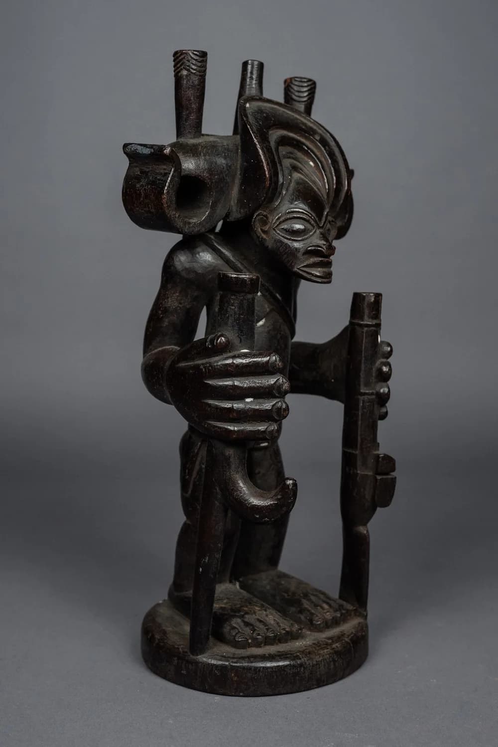 Profile view of a dark wood African statue holding carved pipes, against a grey background
