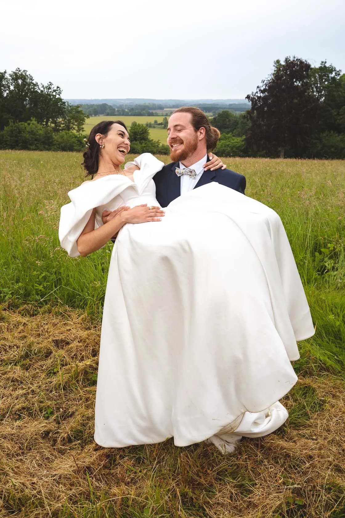 Le marié porte la mariée dans ses bras, tous deux éclatent de rire, robe blanche lumineuse dans un cadre champêtre