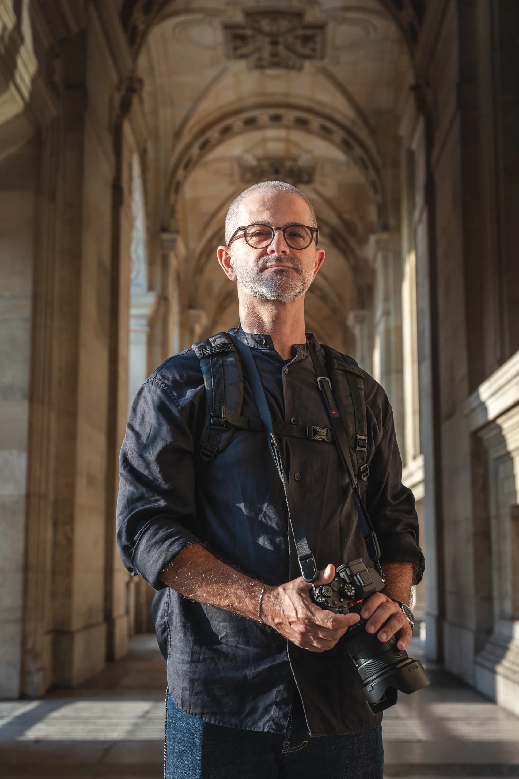Portrait d’un photographe barbu avec appareil photo et sac, dans une colonnade du Louvre